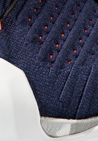 Modrá sportovní bota s texturovaným svrškem z látky, který obsahuje malé oranžové perforace, kontrastní bílou gumovou podrážku a štíhlý, moderní design.