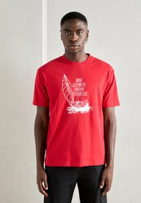 Red cotton t-shirt featuring a white graphic print of a sailboat and text. Short sleeves, round collar, and relaxed fit.
