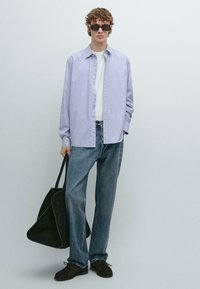 Camicia a righe lilla chiaro con bottoni, t-shirt bianca, jeans blu, scarpe di camoscio nere e borsa tote nera. Il modello ha capelli ricci e occhiali da sole.