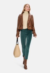 Brown leather cropped jacket, beige turtleneck, teal corduroy pants, brown heeled boots, and holding a beige tote bag with black accents.