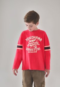 Rød langærmet t-shirt med stort hvidt tryk med teksten "YOKOHAMA" og "DRAGSTERS", sorte striber på ærmerne og en afslappet pasform.
