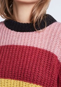 Donna che indossa un maglione di lana con colletto nero e ampie righe orizzontali rosa, rosse e gialle, con capelli castani chiari visibili.