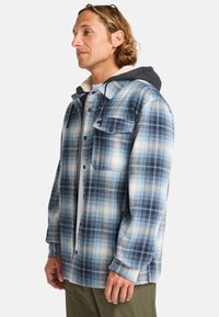 Flannel jacket in blue and white plaid pattern with a soft, textured interior, button closure, and a hood. Notable patch pocket on chest.