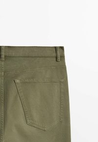 Poche arrière et passants de ceinture d’un pantalon vert olive sur fond blanc, mettant en avant les détails de couture et la texture du tissu.