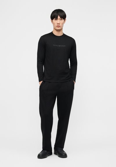 Νέος άνδρας που στέκεται με τα χέρια στις τσέπες, φοράει μαύρο μακρυμάνικο πουκάμισο, μαύρο παντελόνι και μαύρα αθλητικά παπούτσια, μπροστά από απλό ανοιχτόχρωμο φόντο.