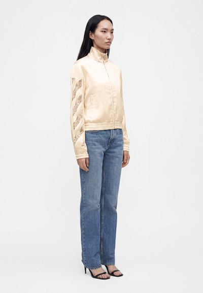Femme debout portant une veste en satin beige avec des bandes en dentelle aux manches, un jean droit bleu et des sandales à talons noires sur fond blanc.