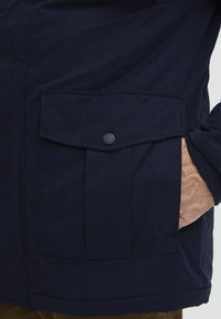 Navy blue jacket featuring a large flap pocket with a black button, textured fabric, and a fitted cuff with a ribbed edge.