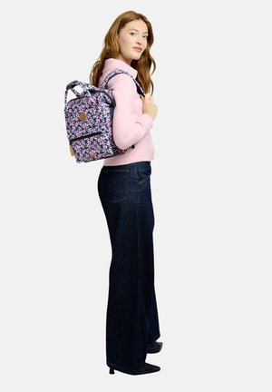 Junge Frau, die einen pinkfarbenen Pullover und dunkle Jeans trägt, mit einem kleinen Rucksack mit Blumenmuster über einer Schulter, seitlich stehend.