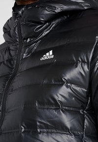 Doudoune noire avec une texture brillante et matelassée. Dispose d'une fermeture éclair et d'un logo Adidas blanc brodé sur la poitrine.