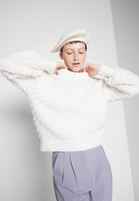 Maglione bianco lavorato a maglia con orlo a costine e motivo testurizzato, indossato con pantaloni grigi plissettati e un berretto coordinato.