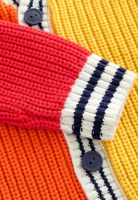 Mehrfarbige Strickjacke mit roten, orangefarbenen, gelben und weißen Abschnitten, ergänzt durch navyblaue Streifenakzente und dunkle Knopfdetails.