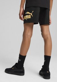 Pantalones cortos negros con el logo dorado de Puma, un ajuste cómodo, combinados con zapatillas negras y calcetas negras acanaladas, mostrando un aspecto deportivo y informal.