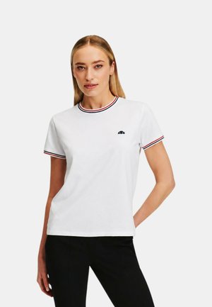 Donna con capelli biondi lisci indossa una t-shirt bianca a maniche corte con rifiniture a righe rosse e nere e piccolo logo nero, abbinata a pantaloni neri.