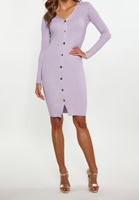 Kvinna klädd i en figurnära lila ribbad stickad klänning med långa ärmar, knäppning fram, sprund i fållen och guld höga klackar.