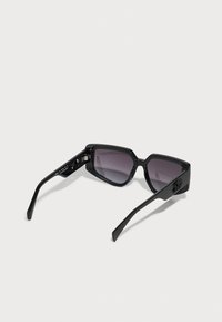 Gafas de sol negras con diseño geométrico, lentes oscuros con degradado, montura de plástico suave y brazos delgados. Acentos metálicos visibles en las patillas.
