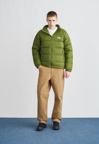 Zaļa pūdera jaka ar izšuves dizainu, rāvējslēdzēja kabatām un logotipu uz krūtīm, kombinēta ar bēšām biksēm un melniem apaviem uz zilas fona.