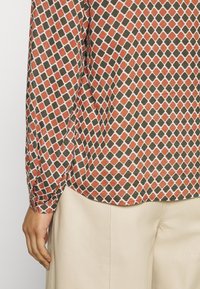 Chemise à manches longues avec un motif géométrique en orange, olive et blanc sur un tissu texturé. Présente un ourlet arrondi et des poignets de manches froncés.