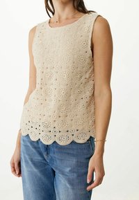 Débardeur en crochet beige avec motifs circulaires, ourlet festonné et design sans manches, associé à un jean bleu clair.