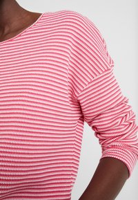 Rot-weißer gestreifter Strickoberteil mit weitem Halsausschnitt und langen, gerafften Ärmeln. Der Stoff hat eine strukturierte Oberfläche, die die visuelle Tiefe verstärkt.