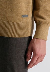 Lichtbruine gebreide trui met geribde zoom en manchetten; heeft aan de zijkant een klein metalen label met de tekst "pierre cardin".