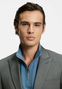 Grå tekstureret habitjakke over en lyseblå trøje med lynlås i halsen. Jakken har en hakrevers og en brystlomme. Enkel design uden mønstre.