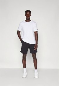 Hvid T-shirt med korte ærmer, sorte shorts med logo, hvide sokker og hvide sneakers. Modellen står foran en neutral baggrund.