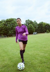 Lila hosszú ujjú focimez fekete díszítésekkel, fekete rövidnadrág és térdig érő fekete zokni. Egy focilabda van a lábánál a zöld pályán.