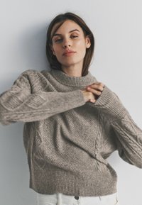 Gebreide beige trui met een hoge geribde kraag, een gestructureerd lijfje en kabelgebreide mouwen, gedragen over een witte broek.