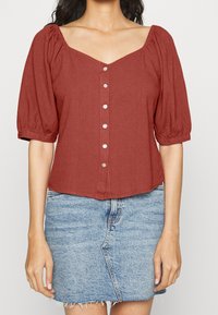 Rustfarbige Bluse mit kurzen Ärmeln, V-Ausschnitt und puffigen Ärmeln. Verfügt über eine Knopfverschluss vorne und einen cropped Schnitt.