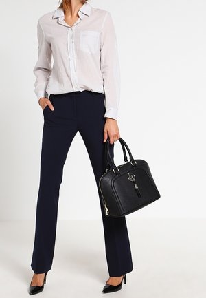 Borsa nera dalla forma arrotondata e superficie sagomata, con accessori in argento e decorazione a nappa. Abbinata a una camicia chiara e pantaloni blu scuro.