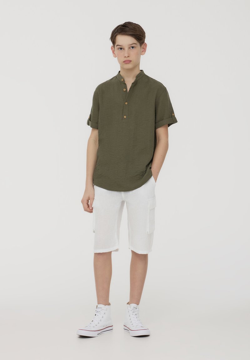 Camicia verde oliva a maniche corte con bottoni, abbinata a pantaloni cargo bianchi. Il modello indossa sneakers alte bianche.
