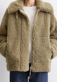 Beige texturerad sherpa-jacka med stor krage, framzipper och tryckknappar, bärs över en vit ribbad tröja och blå jeans.