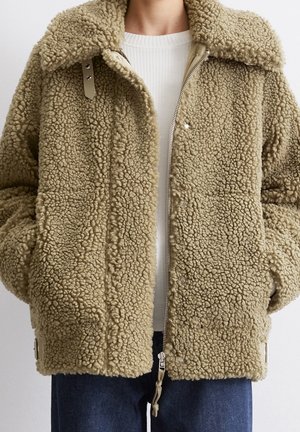 Veste en sherpa beige texturée avec grand col, fermeture éclair à l'avant et boutons-pression, portée sur un pull en maille blanche et un jean bleu.