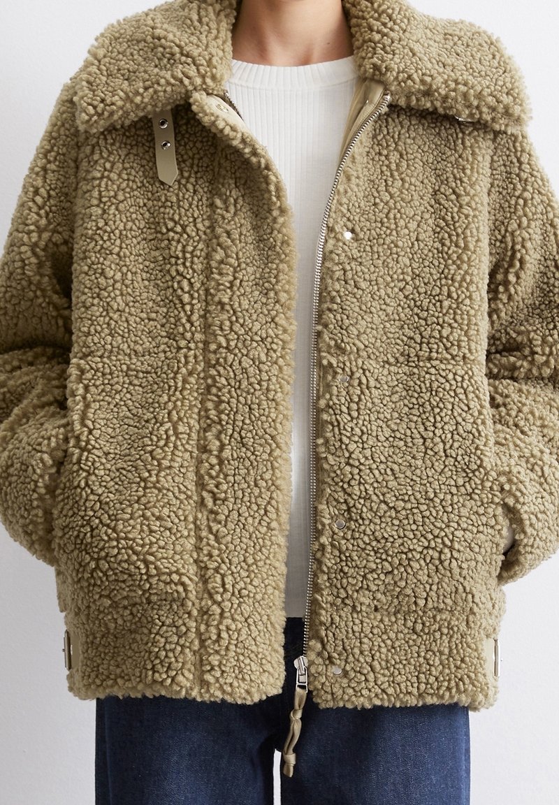 Beige texturerad sherpa-jacka med stor krage, framzipper och tryckknappar, bärs över en vit ribbad tröja och blå jeans.