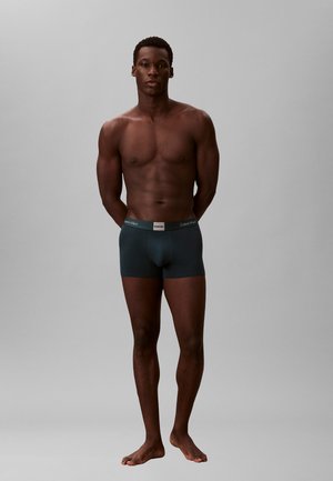 Mannelijk model staat op blote voeten met armen achter de rug, draagt donkerblauwe Calvin Klein boxershorts tegen een effen grijze achtergrond.