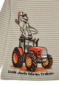 T-shirt grigia a righe con un dinosauro dei cartoni animati su un trattore rosso, con ruote nere, che fa il segno della pace e indossa un cappellino e scarpe da ginnastica.