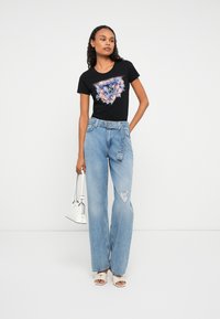 Kvinne iført svart floral grafisk T-skjorte, høytlivte lyseblå vide jeans med belte, hvite hæl-sandaler, og holder en hvit håndveske.