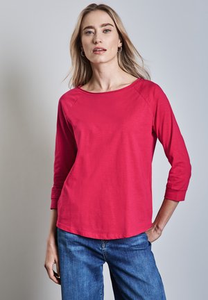 Fuchsia langarmtop met een wijd decolleté en driekwart raglanmouwen, gecombineerd met blauwe spijkerbroek. Soepele textuur en casual pasvorm.