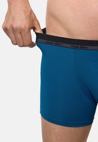 Tyrkysové boxerky s černým vzorovaným pasem označeným "murder." Materiál působí hladce, přiléhavě a pružně, poskytující těsné střih.