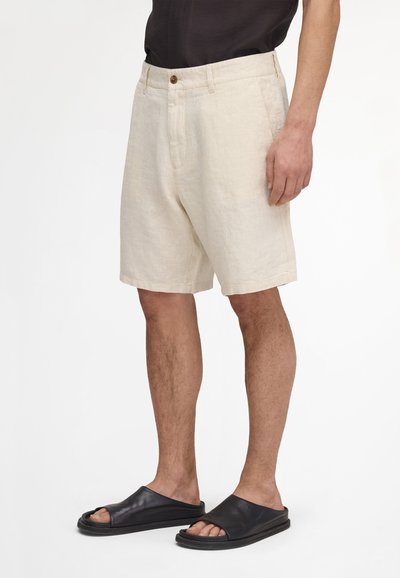 Shorts beige jusqu'aux genoux avec bouton à l'avant et poches, portés par une personne avec des sandales noires et une chemise noire, debout sur un fond blanc.