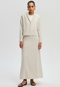 Beige stickad jacka med en kortad design och knäppning, i kombination med en matchande långkjol. Båda har strukturerade fållar och detaljer.