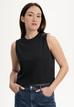 Femme aux longs cheveux bruns portant un haut court noir sans manches et un jean bleu, debout les mains dans les poches sur fond blanc.