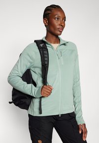 Hellgrüne Zip-Jacke aus strukturiertem Stoff, hoher Kragen und Reißverschlusstaschen, kombiniert mit schwarzen Cargo-Hosen und einem schwarzen Rucksack.