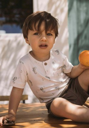 Unge dreng iført skjorte og shorts med søhestetryk, siddende udendørs og holder et orange frugt i den ene hånd og en legetøjsbil i den anden.