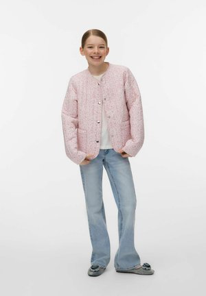 Smilende pige med håret bundet tilbage iført en lyserød blomsterprintet jakke med knapper, hvid skjorte, lyseblå jeans og grå hjemmesko.