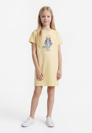 Ung pige med langt blondt hår iført en gul kjole med en bjørnegrafik, der holder en kurv, hvide sneakers, stående mod en hvid baggrund.