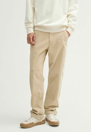 Beige cotton pants with a relaxed fit, textured material, subtle stitching details, and side pockets, paired with cream sneakers.
