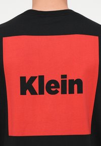 Crna pamučna majica sa hrabrim crvenim pravokutnim naglaskom na kojem se nazad nalazi riječ "Klein" u velikom crnom fontu.