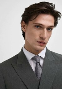 Abito grigio su misura con revers a punta, camicia bianca e cravatta grigia strutturata. Tessuto liscio con una leggera lucentezza. Ritratto ravvicinato.