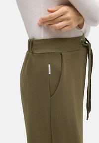 Pantalones de color verde oliva con cintura ajustable, bolsillos laterales y una pequeña etiqueta de marca. Tejido suave con un ajuste entallado.